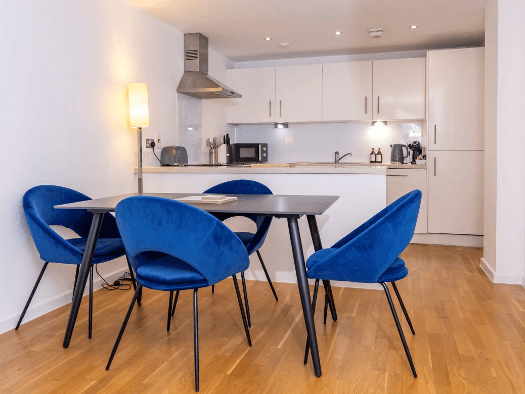 Bright dining area with round table, blue accent chairs, and fully fitted white kitchen at The Sycamore Suite, Enterprise Place, Woking Bright dining area with round table, blue accent chairs, and fully fitted white kitchen at The Sycamore Suite, Enterprise Place, Woking
