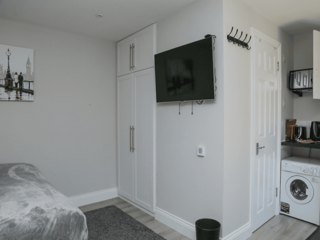 Wall-mounted flat-screen TV, wardrobe, and kitchen view in compact serviced studio – Crofton Park, London Wall-mounted flat-screen TV, wardrobe, and kitchen view in compact serviced studio – Crofton Park, London