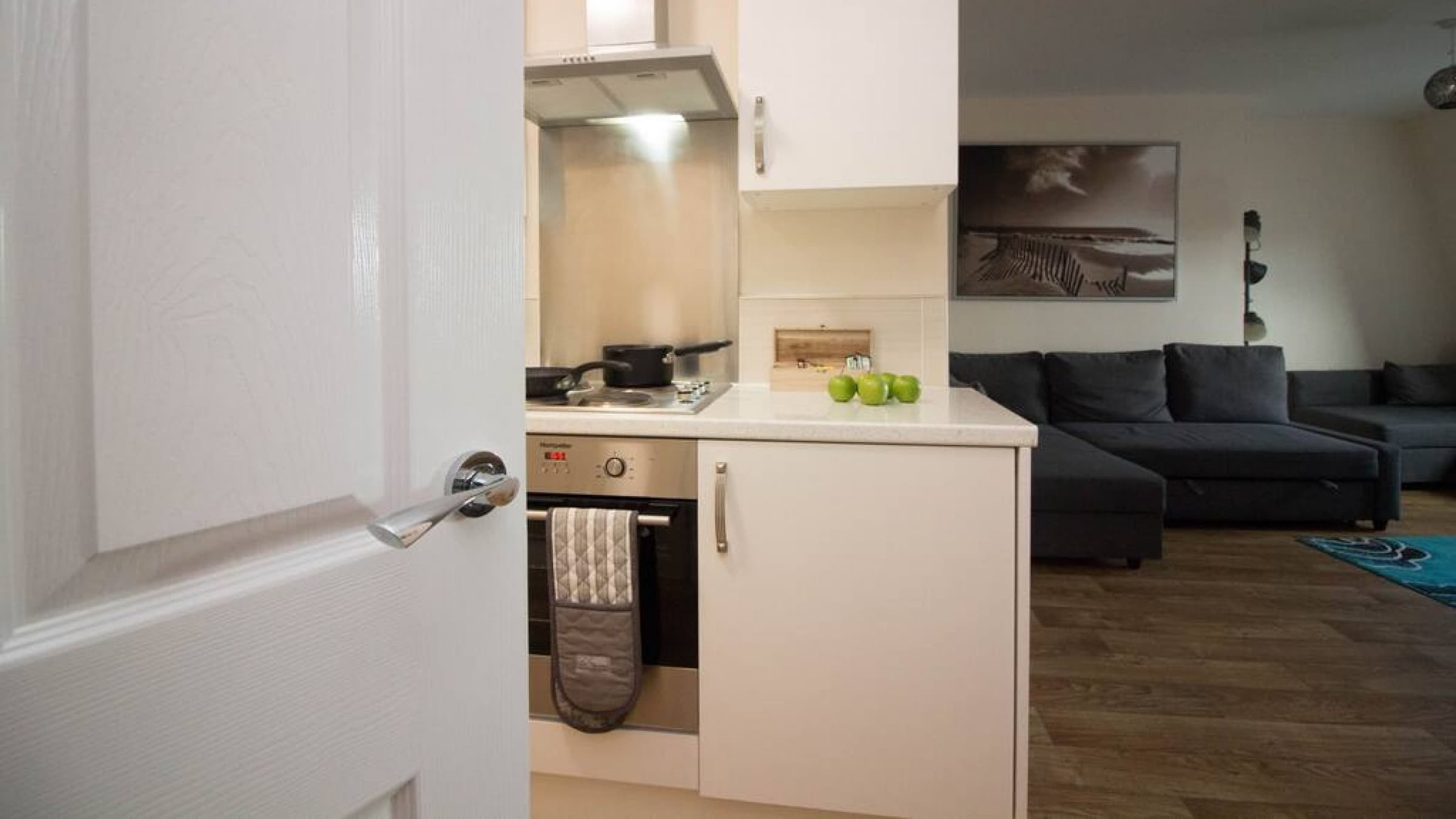 Open-plan kitchen with integrated oven, cooktop, and lounge view toward grey sofas – Castle 18 - Southampton Open-plan kitchen with integrated oven, cooktop, and lounge view toward grey sofas – Castle 18 - Southampton