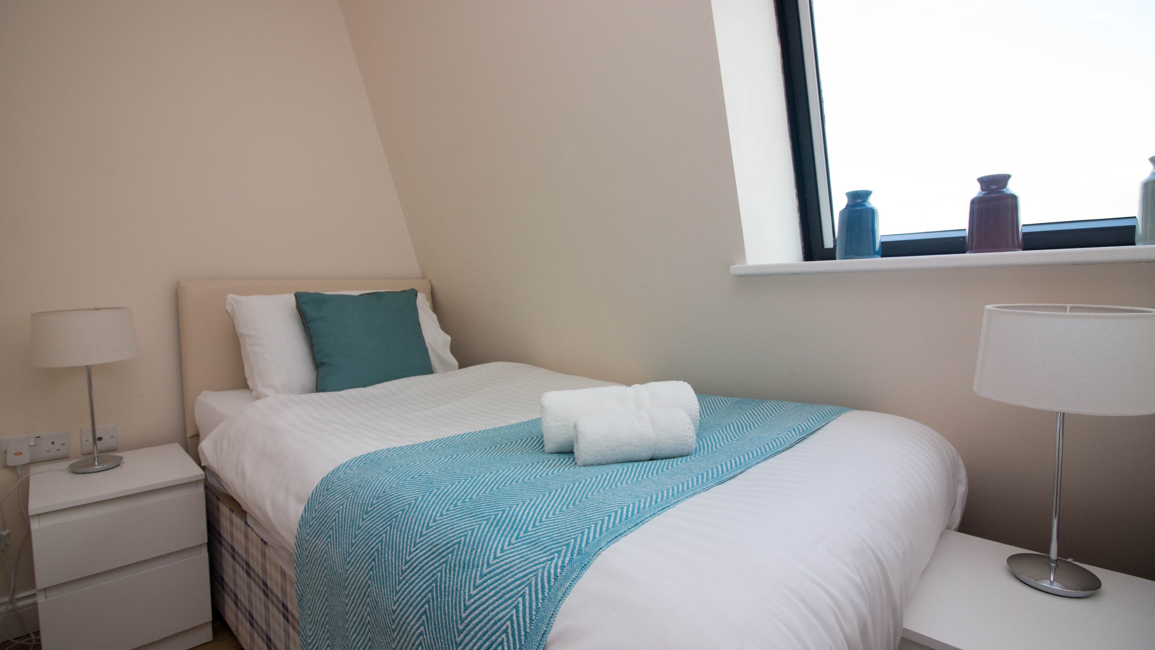 Single bedroom with blue throw, bedside lamp, and bright window in modern serviced apartment – Castle 19 - Southampton Single bedroom with blue throw, bedside lamp, and bright window in modern serviced apartment – Castle 19 - Southampton