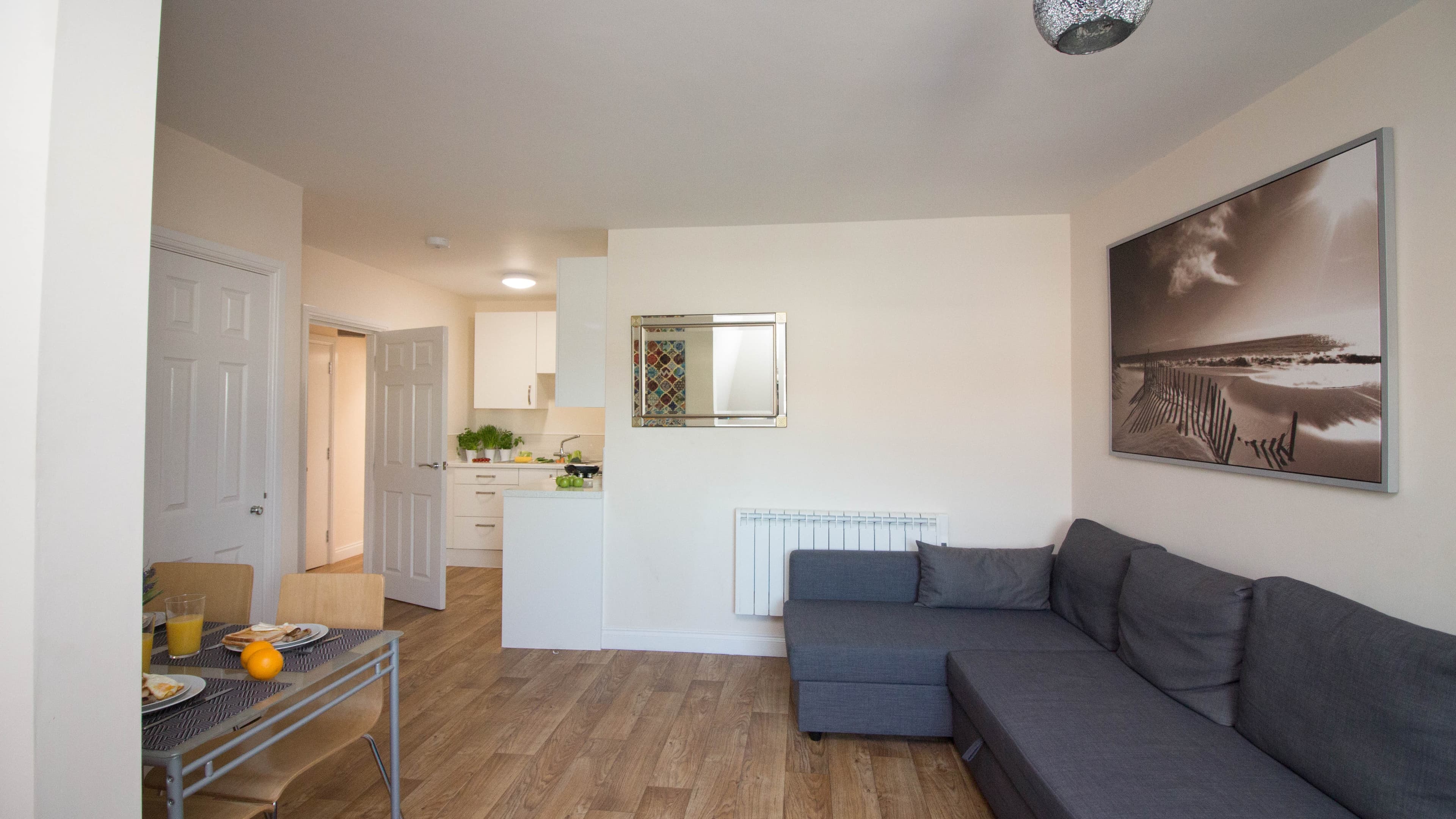 Open-plan living room and kitchen with corner sofa, wood flooring, wall art, and hallway access – Castle 19 - Southampton Open-plan living room and kitchen with corner sofa, wood flooring, wall art, and hallway access – Castle 19 - Southampton