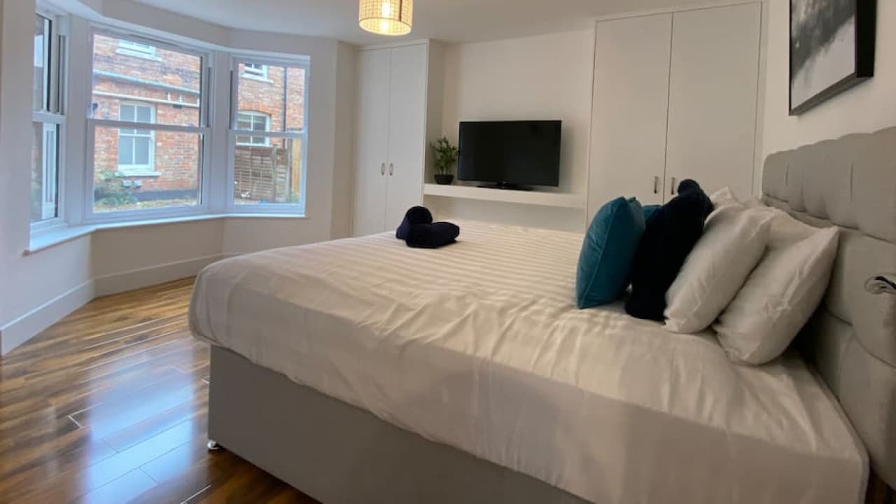 Spacious bedroom with double bed, blue cushions, grey headboard, window view and TV – De Parys Flat 2, Bedford Spacious bedroom with double bed, blue cushions, grey headboard, window view and TV – De Parys Flat 2, Bedford