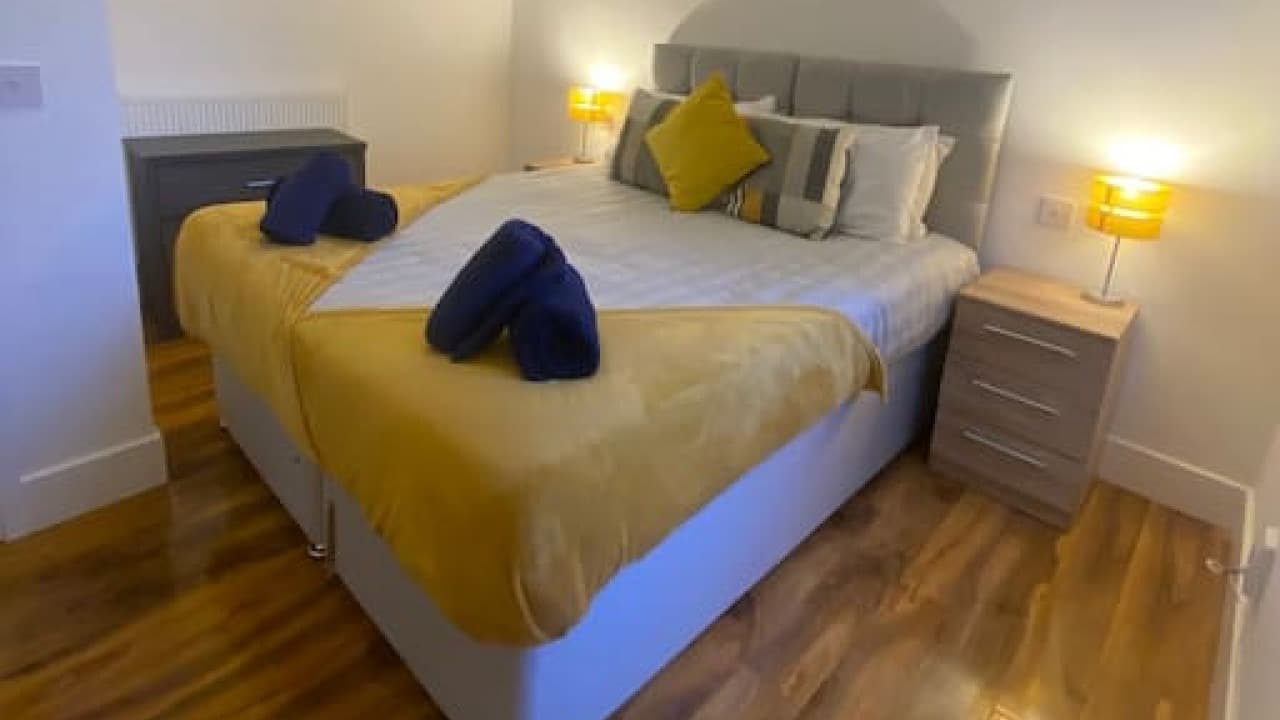 Double bedroom with yellow accent cushion, wood bedside table, lamp, and window light – De Parys Flat 3, Bedford Double bedroom with yellow accent cushion, wood bedside table, lamp, and window light – De Parys Flat 3, Bedford