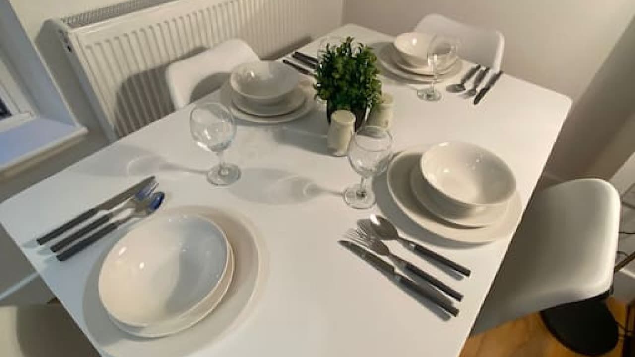 White dining table set for four, potted plant centerpiece, and grey chairs in bright living space – De Parys Flat 4, Bedford White dining table set for four, potted plant centerpiece, and grey chairs in bright living space – De Parys Flat 4, Bedford