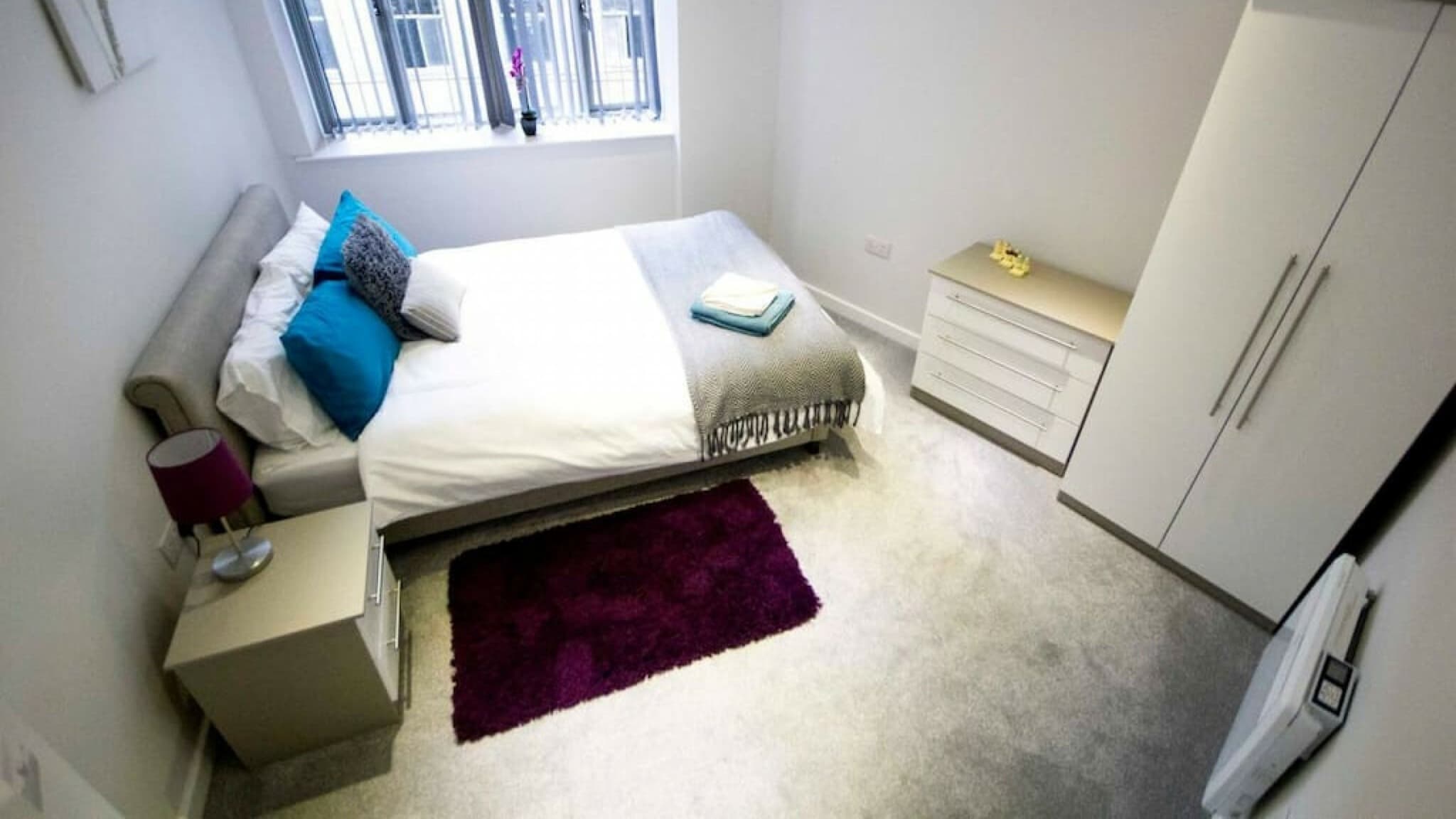 Bright single bedroom with teal pillows, white linens, and purple rug near window and wardrobe – Grattan 2 - Bradford Bright single bedroom with teal pillows, white linens, and purple rug near window and wardrobe – Grattan 2 - Bradford